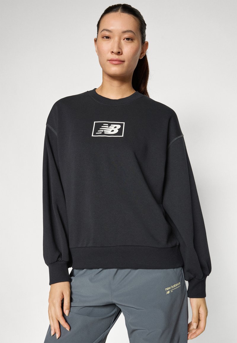 Felpa nera con logo bianco di New Balance sul petto, vestibilità oversized, polsini a coste e scollatura tonda. Abbinata a pantaloni grigi.