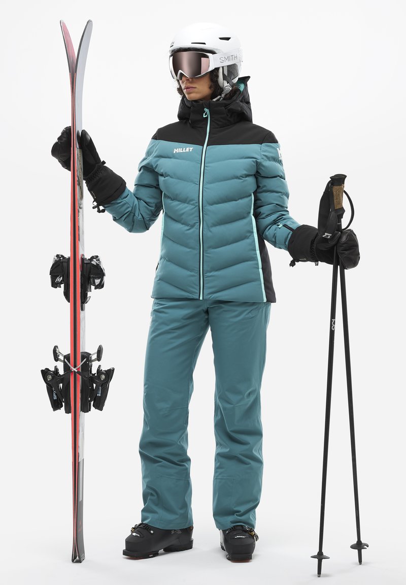Veste de ski rembourrée teal avec des accents noirs, associée à un pantalon assorti. Tient des skis et des bâtons. Les accessoires comprennent un casque blanc et des lunettes de ski.