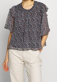Bluse med blomsterprint i gennemsigtig navy stof med flæse korte ærmer, rund halsudskæring og plisseret front. Båret med lyse bukser.