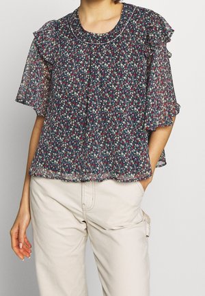 Bluse mit Blumenmuster aus durchsichtigem marineblauen Stoff, mit kurzärmeligen Rüschen, runden Ausschnitt und gefältelter Vorderseite. Kombiniert mit hellen Hosen.