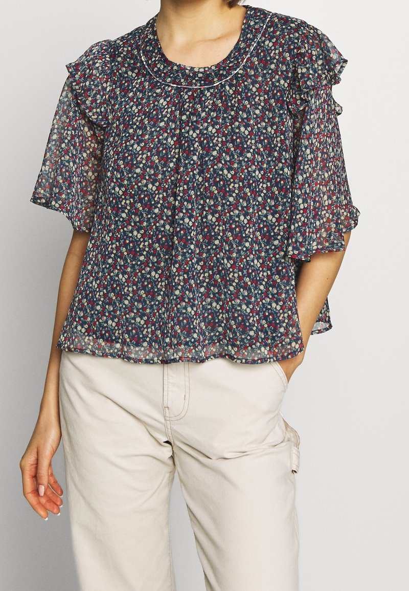 Bluse med blomsterprint i gennemsigtig navy stof med flæse korte ærmer, rund halsudskæring og plisseret front. Båret med lyse bukser.