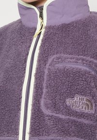 Giacca in pile viola con colletto alto, zip frontale e dettagli color crema. Presenta una tasca sul petto con zip e un dettagliato logo.