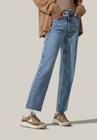 Jeans en denim bleu clair avec une coupe droite, associées à des baskets marron avec des accents blancs et une semelle épaisse beige. Cardigan marron porté ouvert.