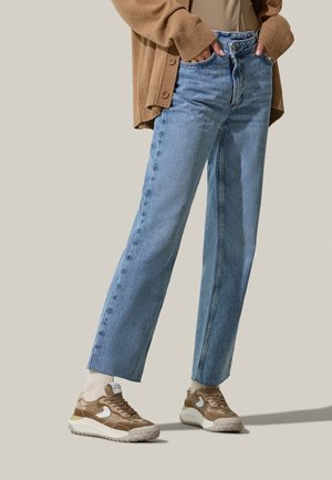 Jeans in denim chiaro con taglio dritto, abbinati a sneakers marroni con dettagli bianchi e suola spessa beige. Cardigan marrone indossato aperto.