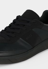 Gros plan d'une chaussure en cuir et daim noir avec des lacets noirs et le nom de la marque Boss embossé sur le côté et la languette.