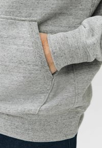 Hanorac gri dintr-un material texturat, cu manșete ribbate și buzunar tip kangaroo. Dispune de un finisaj neted și o croială casual.