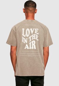 Lys brun oversized t-skjorte med rund hals, med stor hvit tekst "LOVE IN THE AIR" på baksiden og små tekstdetaljer nedenfor.