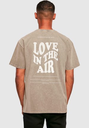 Lys brun oversized t-skjorte med rund hals, med stor hvit tekst "LOVE IN THE AIR" på baksiden og små tekstdetaljer nedenfor.