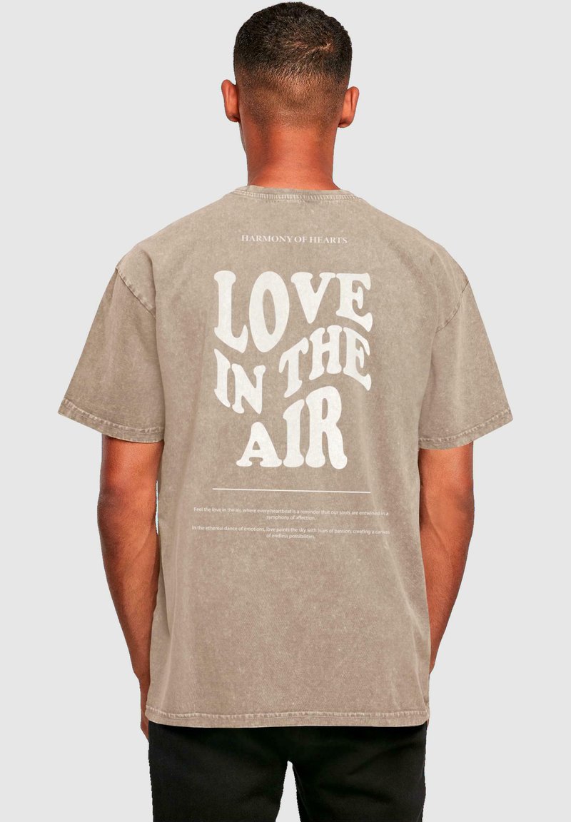 Lys brun oversized t-skjorte med rund hals, med stor hvit tekst "LOVE IN THE AIR" på baksiden og små tekstdetaljer nedenfor.