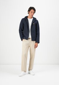 Navy lightweight jacket with a hood, front zip, and yellow logo. Paired with cream chinos and white sneakers for a casual look.