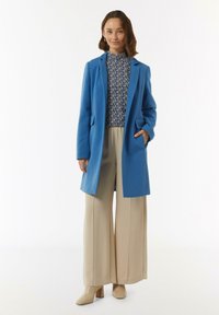 Blauwe lange jas met een gestructureerd ontwerp, beige wijde broek en een gestreept top. De jas heeft zijzakken en een losse pasvorm.