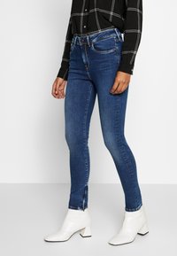 Tunna blå denimjeans med en mjuk textur, subtila blekningar och ankellås, tillsammans med en svart rutmönstrad skjorta och vita klackstövlar.