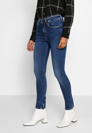 Jeans de mezclilla azules ajustados con una textura suave, un ligero desgaste y cremalleras en los tobillos, combinados con una camisa a cuadros negra y botas de tacón blancas.