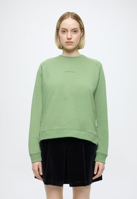 Sweatshirt verde com decote redondo e punhos canelados, apresentando um logótipo aplicado no centro e uma etiqueta na lateral. Combina com uma saia de veludo cotelê preta.