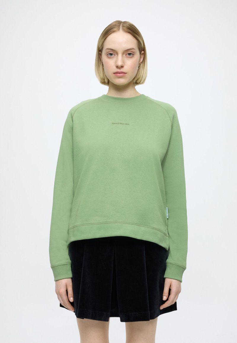 Sweatshirt verde com decote redondo e punhos canelados, apresentando um logótipo aplicado no centro e uma etiqueta na lateral. Combina com uma saia de veludo cotelê preta.