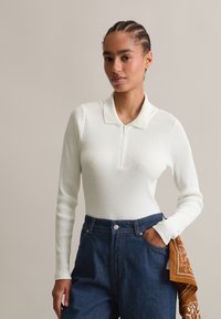 Vrouw met gevlochten haar draagt een witte geribbelde longsleeve polo en hoge blauwe jeans met een oranje patroon sjaal in haar zak gestoken.