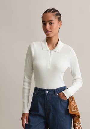 Eine Frau mit geflochtenem Haar trägt ein weißes, geripptes Langarm-Polo und hoch taillierte blaue Jeans, mit einem orangefarbenen, gemusterten Schal, der in die Tasche gesteckt ist.