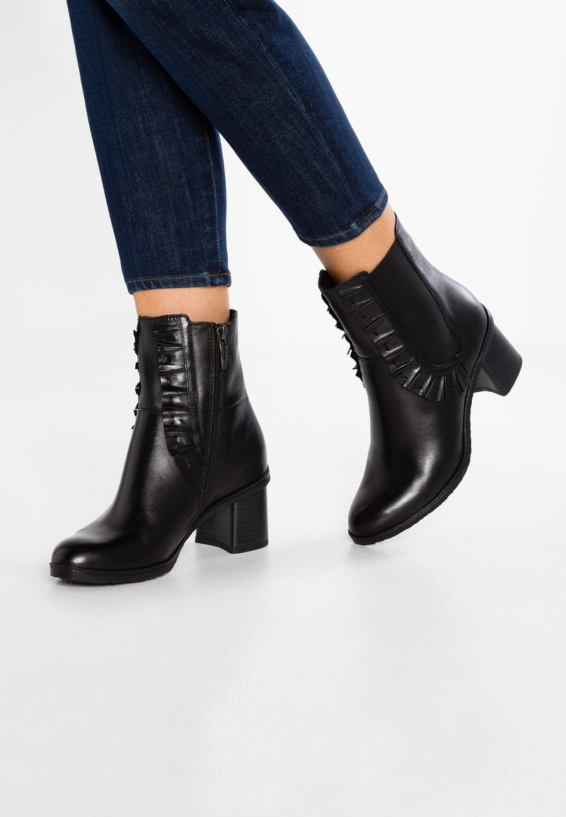 Jambes en jean bleu foncé portant des bottines en cuir noir avec talons blocs bas et fermeture éclair latérale, debout sur un sol blanc.