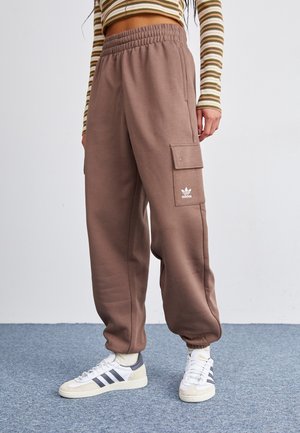 Persoon draagt bruine Adidas cargo joggingbroek, witte sokken, witte sneakers met zwarte strepen en een cropped gestreept shirt met lange mouwen.