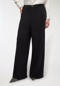 Pantaloni neri a gamba larga con una texture liscia, dotati di chiusura con bottone e tasche frontali. Abbinati a scarpe con punte rifinite in metallo.