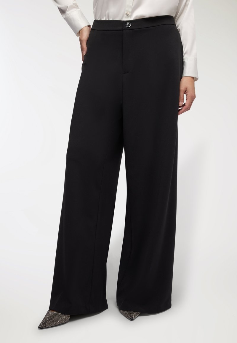 Pantaloni neri a gamba larga con una texture liscia, dotati di chiusura con bottone e tasche frontali. Abbinati a scarpe con punte rifinite in metallo.