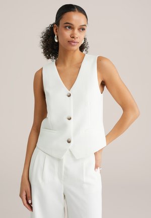 Femme aux cheveux bouclés portant un gilet blanc sans manches à boutons et un pantalon blanc assorti, posant avec une main dans la poche devant un fond uni.