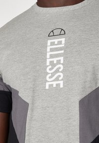 Grå t-shirt med rund halsringning som har vertikal vit text "ELLESSE" och mörkgrå geometriska detaljer på ärmarna och nedre delen.