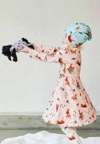 Vaaleanpunainen mekko, jossa on eläinkuosetteja, pitkät hihat ja liehuva hame. Lapsi, joka pitää päässään vaalean sinistä kuviollista pipoa ja pidättelee pehmolelua.