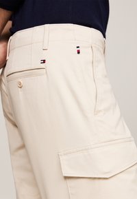 Pantalons cargo beiges avec une texture lisse, dotés d'une poche arrière, d'une fermeture à bouton et d'un logo de marque brodé en accents rouge, blanc et bleu.
