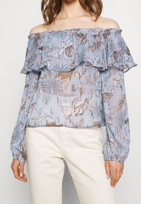 Blouse bleu clair à épaules dénudées avec des couches à volants, présentant des motifs floraux et de feuilles en brun avec des accents métalliques, tissu transparent, manches longues.