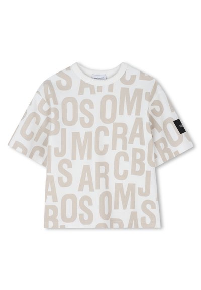 T-shirt blanc à manches courtes avec de grandes lettres beige superposées sur toute la surface, comportant une étiquette en tissu noire sur la manche droite.
