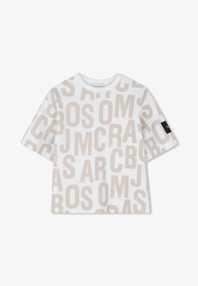 T-shirt blanc à manches courtes avec de grandes lettres beige superposées sur toute la surface, comportant une étiquette en tissu noire sur la manche droite.