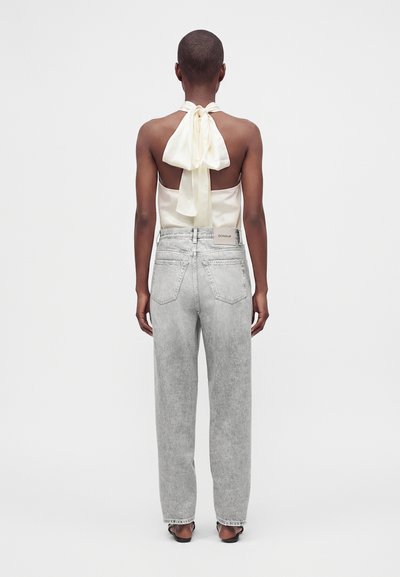 Personne vue de dos portant un haut sans manches crème noué en nœud et un jean taille haute gris clair avec des sandales noires.