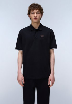 Jonge man met krullend haar, gekleed in een zwart poloshirt met een klein rond logo op de borst, staand tegen een eenvoudige lichte achtergrond.