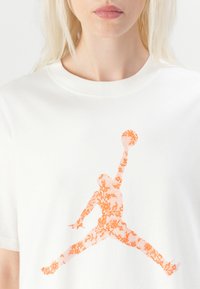 White cotton T-shirt featuring an orange floral-patterned graphic of a jumping figure, round neckline, short sleeves, and relaxed fit.
