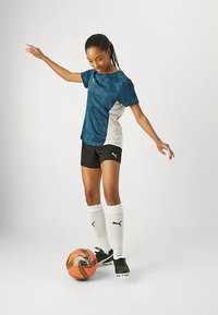 Σετ ποδοσφαίρου Puma με μπλε φανέλα γεωμετρικό μοτίβο, μαύρο σορτς, λευκές κάλτσες μέχρι το γόνατο και μαύρα ποδοσφαιρικά παπούτσια. Πλησίον υπάρχει μια πορτοκαλί μπάλα ποδοσφαίρου.