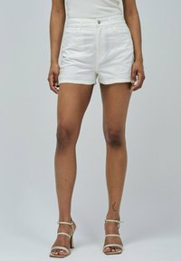 Femme portant des shorts blancs brodés et des sandales à talons hauts blanches, se tenant avec les jambes visibles à partir de la taille devant un fond uni.