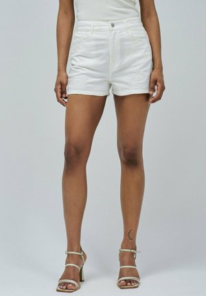 Femme portant des shorts blancs brodés et des sandales à talons hauts blanches, se tenant avec les jambes visibles à partir de la taille devant un fond uni.
