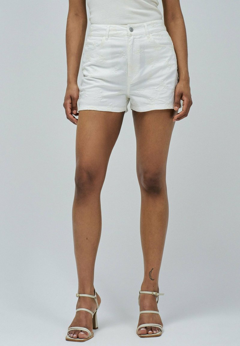 Femme portant des shorts blancs brodés et des sandales à talons hauts blanches, se tenant avec les jambes visibles à partir de la taille devant un fond uni.