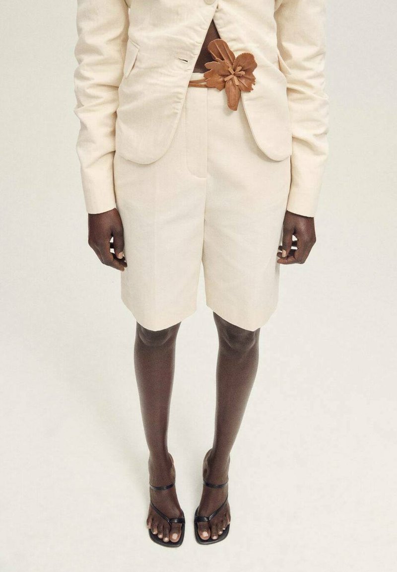 Costume beige avec un blazer sur mesure et un short assorti. Ceinture florale marron en accent. Tongs noires. Tissu lisse, design structuré.