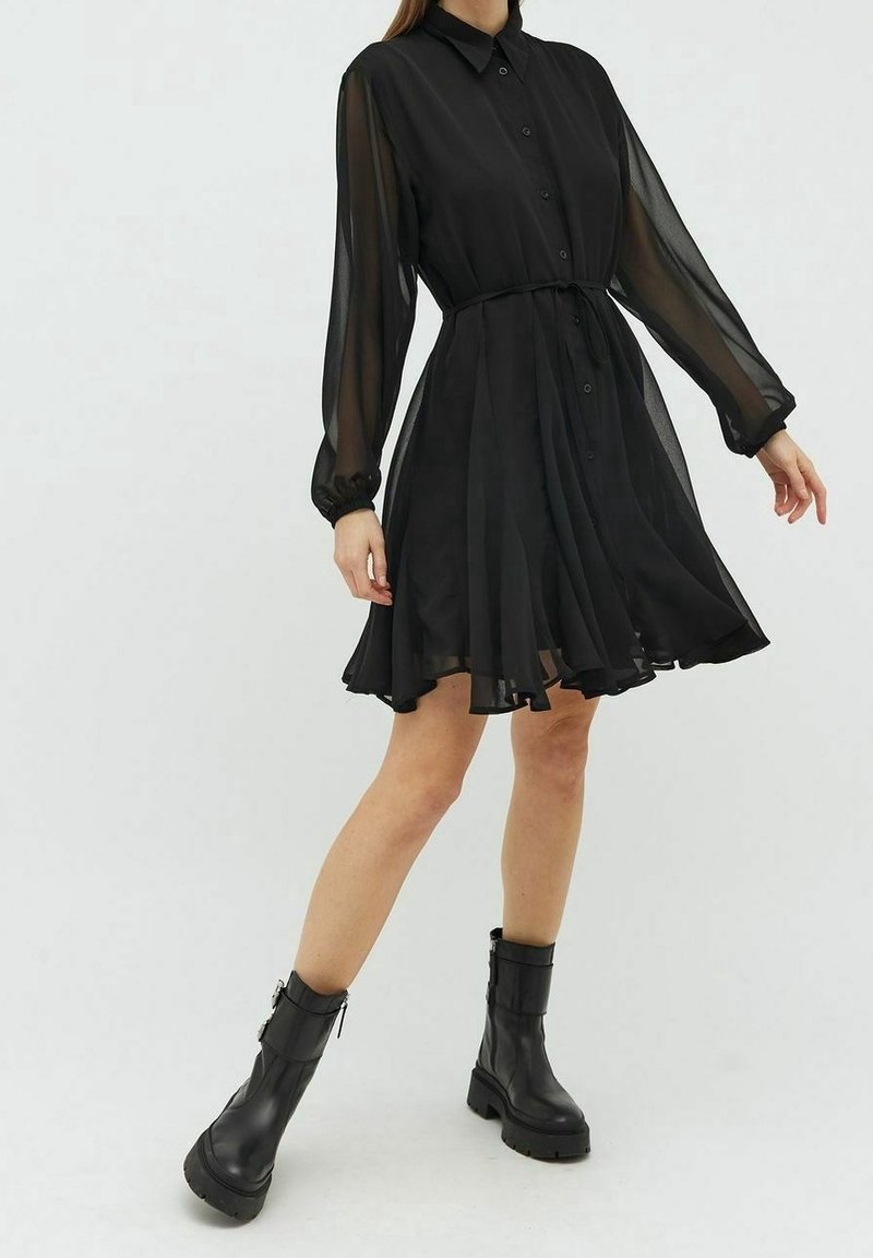 Femme portant une robe noire boutonnée avec des manches longues transparentes et une ceinture à nouer à la taille, associée à des bottines noires à semelles épaisses.