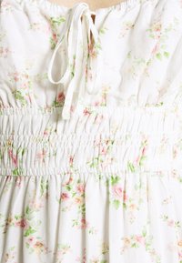 Vestido de tela blanca con estampado floral en rosa y verde, que presenta una cintura elástica fruncida y cintas en el cuello para un ajuste ajustable. Textura suave.