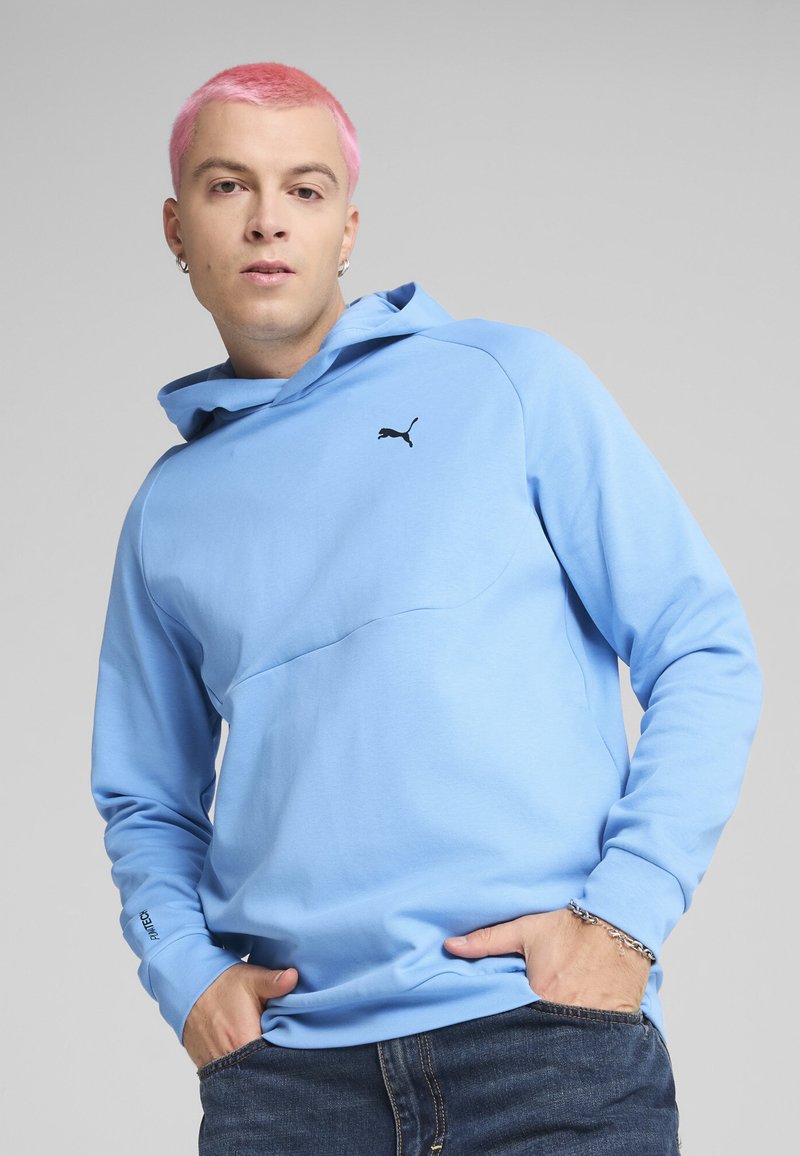 Sudadera azul claro hecha de tejido suave, con un detalle de costura en el frente y el logo negro de Puma en el pecho. Cuenta con un capucho ajustable y mangas largas.
