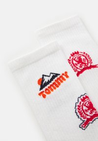 Pair of white ribbed socks featuring embroidered logos: a mountain with orange sun and "Tommy" text, and a red crest design.
