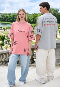 Une jeune femme et un homme se tiennent dehors sur des marches, portant des t-shirts graphiques oversize avec texte et motifs colorés de cœur et de soleil.