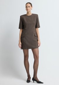 Hnědé strukturované mini šaty s krátkými rukávy, dvojitými zlatými knoflíky a bočními kapsami, doplněné průhlednými punčochami a černými podpatky.