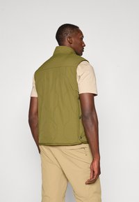Olivgrüne gesteppte Weste mit hohem Kragen, ärmellosem Design und Druckknopfdetails an den Seiten, getragen über einem beigen Hemd und beigen Hosen.