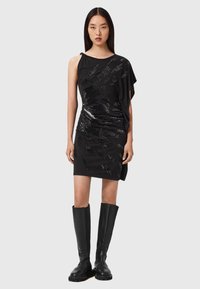 Musta yksishulkkadressi, jossa on teksturoituista paljeteista koristeltu, laskostettu muotoilu ja tiukka siluetti. Yhdistetty polven korkeisiin mustiin saappaisiin.