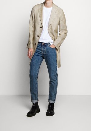 Persona que lleva una blazer beige abotonada sobre una camiseta blanca, jeans azules remangados y botas de cuero negro, con una mano en el bolsillo.