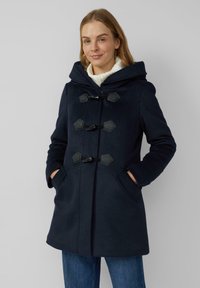 Frau trägt einen marineblauen Mantel mit Knebelverschlüssen und Kapuze über einem weißen Rollkragenpullover und blauen Jeans, die Hände in den Manteltaschen.